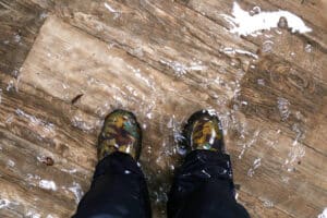 A woman's feet, wearing waterproof rain boots are standing in a flooded house with vinyl wood floors. BEST WATER RESTORATION COMPANY NEAR ME HOLLYWOOD FLORIDA