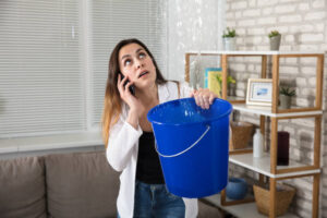 Worried Woman Calling Plumber While Collecting Water Droplets Leaking From Ceiling At Home HOMESTEAD FLORIDA